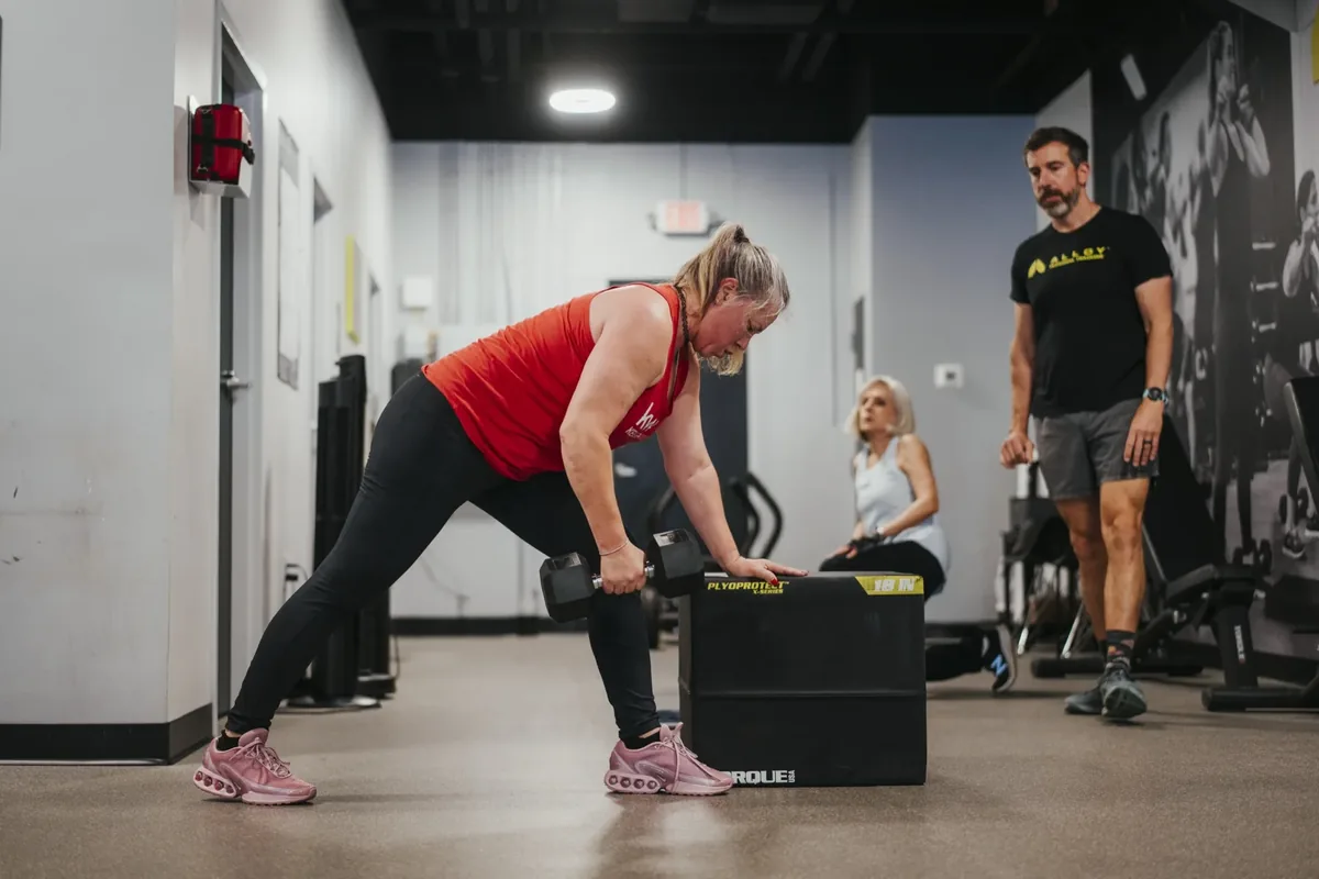 A member performs a heavy single-arm dumbbell row braced on a box while a coach watches her form
