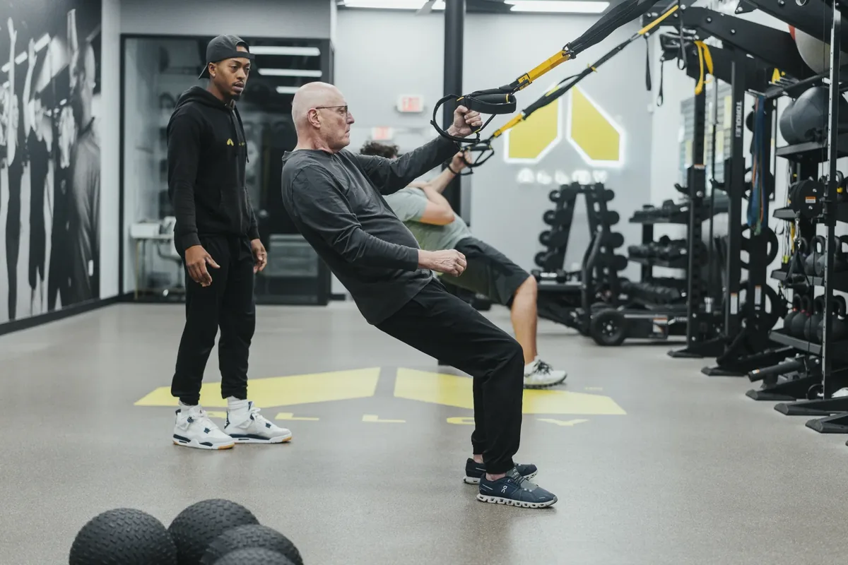 A coach supervises an older member leaning back into a suspension trainer row