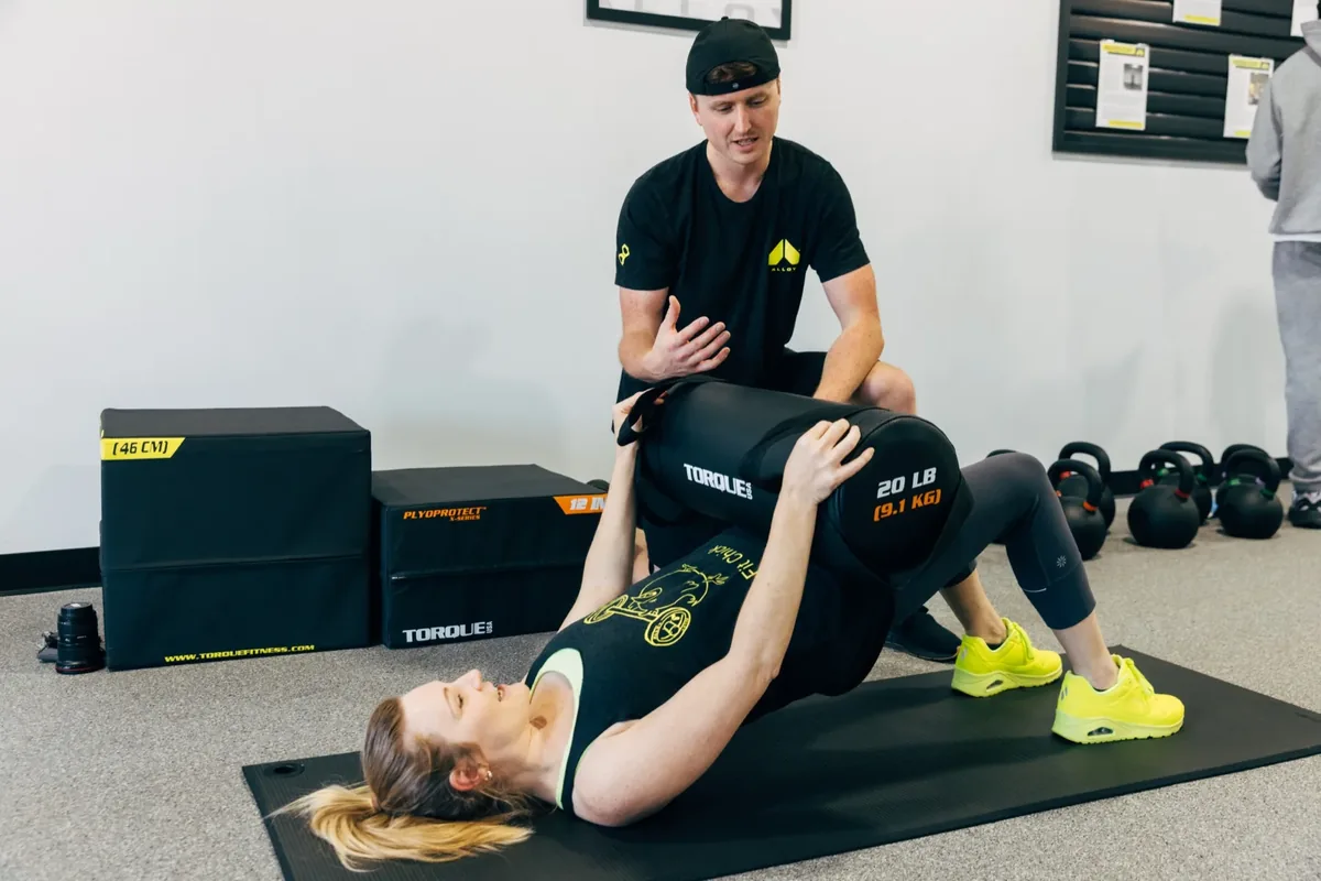 A coach guides a member through a hip bridge on a mat at an Alloy Personal Training studio