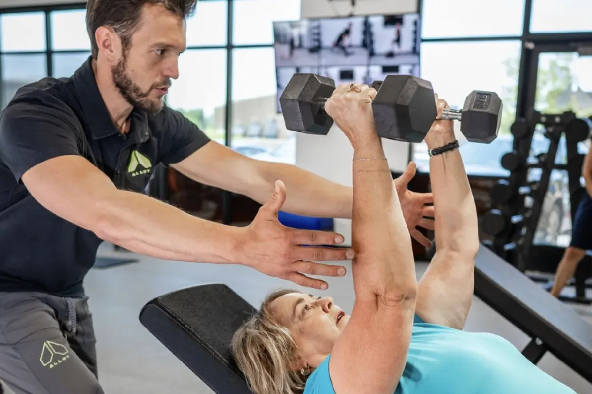 A coach supports a member's arms as she presses dumbbells overhead at an Alloy Personal Training studio